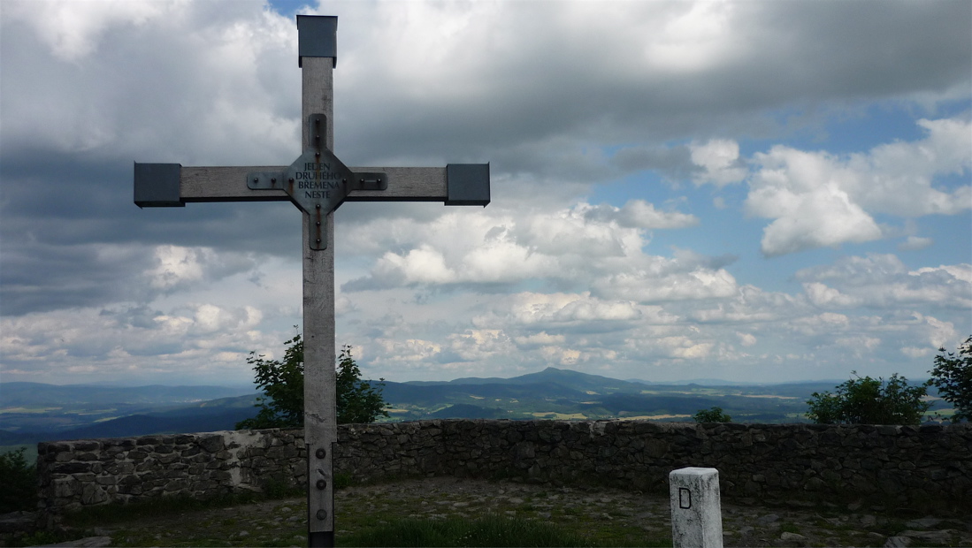 Nápis na kříži: "Jeden druhého břemena neste". V dálce Ještěd.